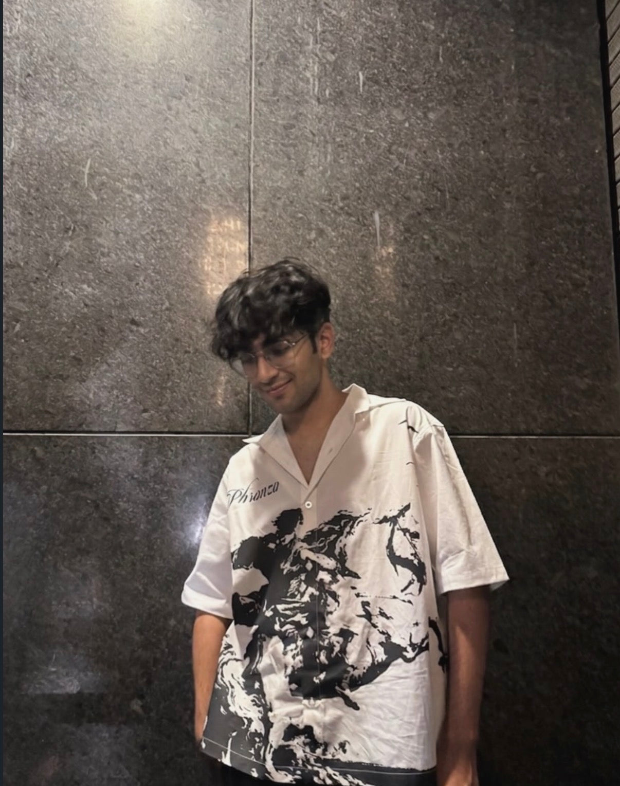 Person wearing a white shirt with black abstract design against a dark tiled wall.